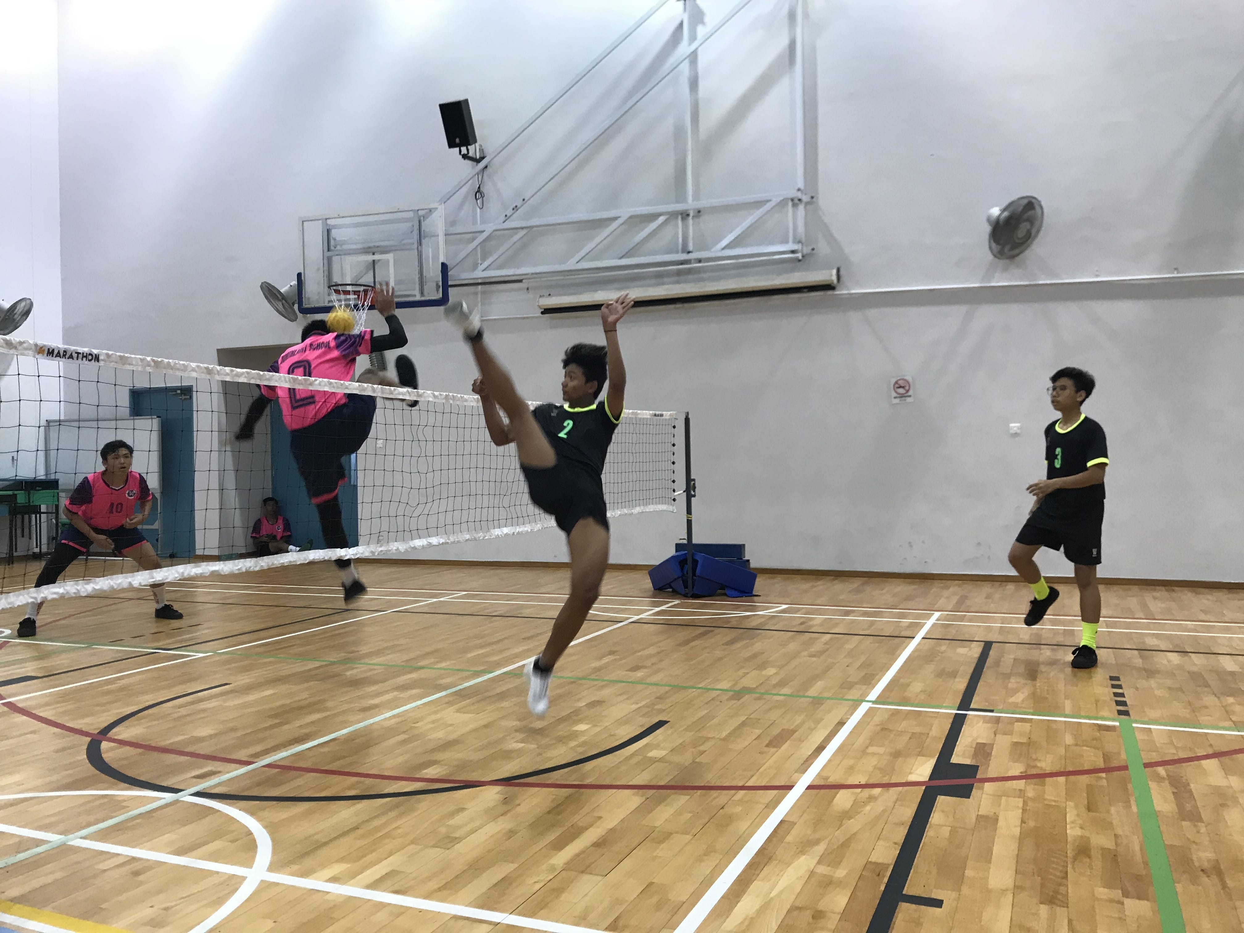 Players in pink and black uniforms compete in a sepak takraw match indoors, with one player kicking over the net.